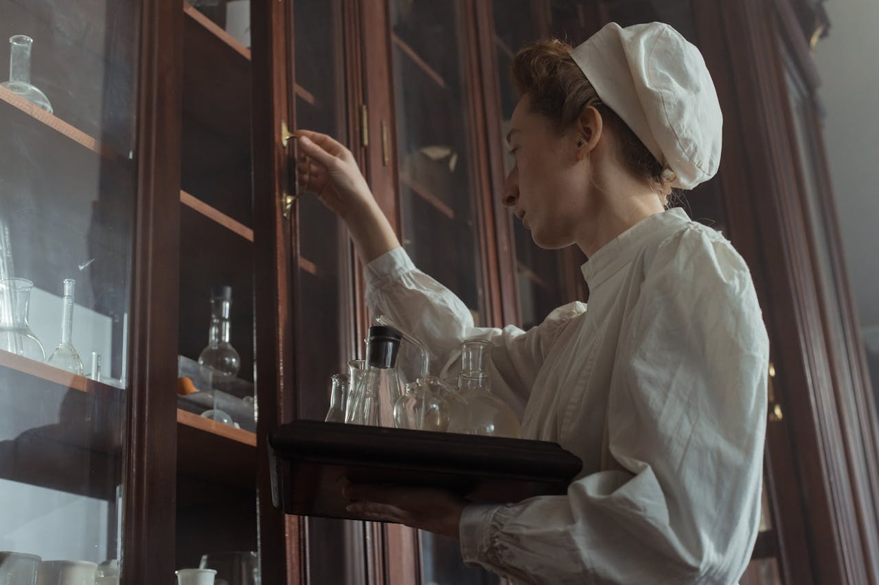 A female lab technician in vintage attire accessing a wooden cabinet in a pharmacy.