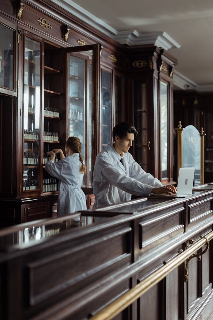 heros-img Pharmacists in a traditional pharmacy setting with wooden cabinetry and a laptop.